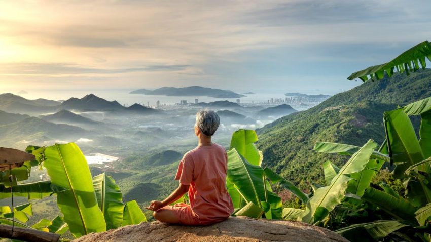 Woman meditating on rock overlooking lush green mountains and city at dawn.